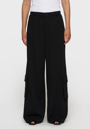 Sorte bukser med vide ben og sidelommer, båret med en hvid ærmeløs top, vist forfra fra talje til fødder.