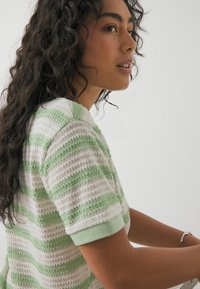 Sieviete ar gariem cirtainiem tumšiem matiem valkā baltu un gaiši zaļu svītrainu adītu īsroku kreklu, skatioties uz sāniem pret vienkāršu fonu.