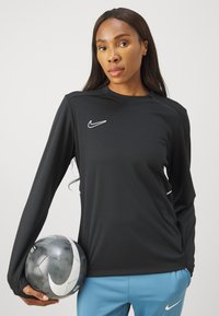 Černé sportovní tričko s dlouhým rukávem a bílým logem Nike, v kombinaci se světle modrými kalhotami. Drží šedý fotbalový míč s černobílými akcenty.
