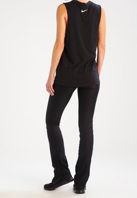 Mujer de pie de espaldas frente a la cámara, vistiendo una camiseta sin mangas negra de Nike, pantalones negros y zapatos negros, contra un fondo blanco.