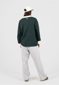 Sudadera oversized verde oscuro con un cuello blanco, combinada con pantalones de pierna ancha grises. El modelo también lleva una gorra blanca con detalles verdes.