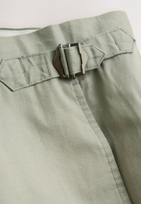 Világoszöld szövetnadrág rövidnadrág fém csattal a deréknál, varrott részletekkel és sima textúrával. Közeli kép.