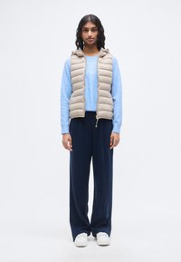 Μπεζ γιλέκο με επένδυση και κουκούλα πάνω από ένα ανοιχτό μπλε πουλόβερ, σε συνδυασμό με σκούρα μπλε παντελόνια με φαρδιά μπατζάκια και λευκά αθλητικά παπούτσια. Απλός σχεδιασμός, ελαφρύ ύφασμα.