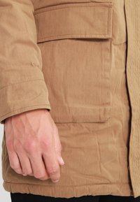 Khaki bawełniana kurtka o teksturowanym wykończeniu, z boczną kieszenią, mankietem i ściąganym dolnym brzegiem.