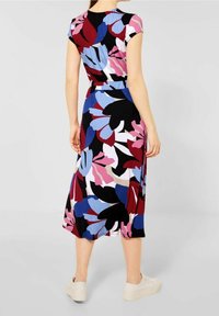 Femme portant une robe midi florale colorée avec des manches courtes, se tenant les mains détendues sur les côtés, associée à des baskets blanches.