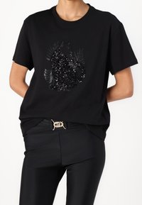 Black t-shirt with short sleeves, featuring a sequin-embellished cat design on the front. Worn with black slim-fit trousers and a belt.