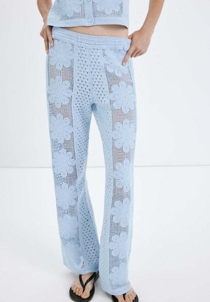 Pantalones de crochet azul claro con patrones florales, combinados con top corto a juego y chanclas negras, manos apoyadas en la cintura.