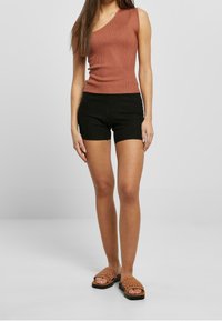 Gestreiftes, einseitiges, rostfarbenes Oberteil kombiniert mit schwarzen, gerippten Shorts. Das Model trägt braune, gewebte Sandalen mit flacher Sohle.