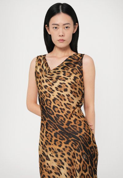 Mulher com cabelo preto liso a usar um vestido a meia altura de leopardo com decote drapeado, de pé com uma mão atrás das costas.