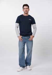 Tumši zils T-krekls ar pelēkām garām piedurknēm zem tā, kombinēts ar gaiši zilām džinsa biksēm un baltām sporta kurpēm. Vienkāršs dizains, brīvs piegriezums.