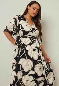 Blommig omslagsklänning i svart och krämvit, med 3/4-långa ärmar med bell-modell, en midjeband och textil med stora blommönster.