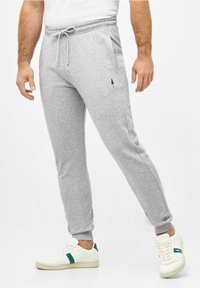 Graue Baumwoll-Jogginghosen mit elastischem Bund, Kordelzug, Seitentaschen und einem kleinen schwarzen Baum-Logo am linken Oberschenkel. Weiße Sneakers mit grünen Akzenten.