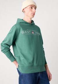 Felpa verde in cotone con tasca a marsupio, presenta un logo bianco e strisce sul petto. Indossata con jeans di denim blu.