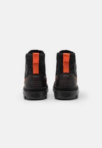 Chaussures montantes noires en tissu texturé, avec languette arrière orange et semelle en caoutchouc noire. Présentent une marque sur le côté avec des détails en relief.