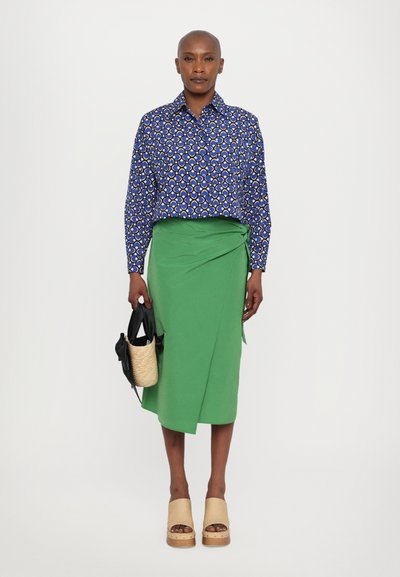 Blue patterned shirt with circular motifs, green wrap skirt, wooden platform shoes, and a black and straw bag.