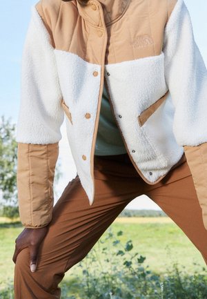 Osoba nosząca beżowo-białą polarową kurtkę z logo The North Face oraz brązowe spodnie, na zewnątrz w pobliżu zieleni i niebieskiego nieba.