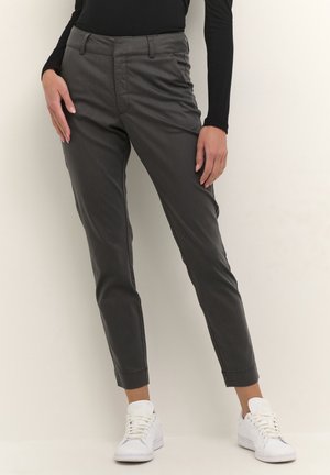 Personne portant un pantalon gris foncé longueur cheville, un haut noir à manches longues et des baskets blanches, debout devant un fond clair et uni.