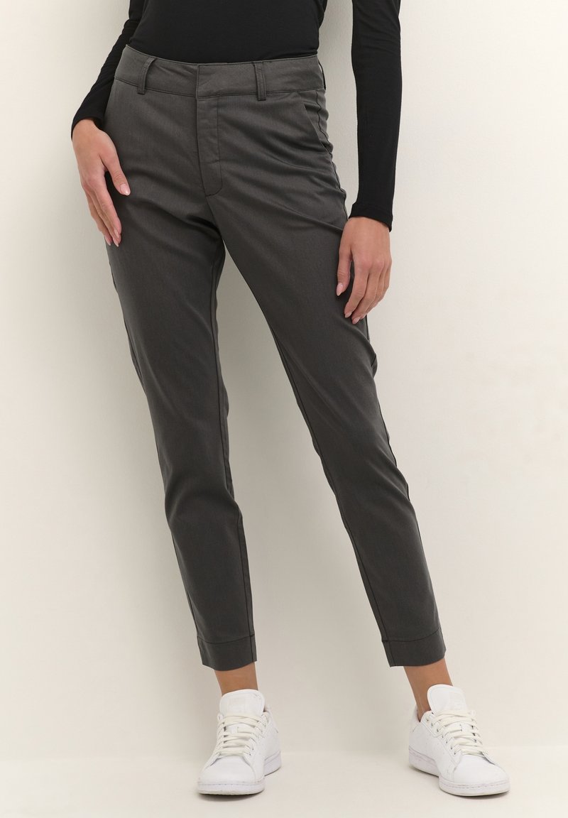 Personne portant un pantalon gris foncé longueur cheville, un haut noir à manches longues et des baskets blanches, debout devant un fond clair et uni.