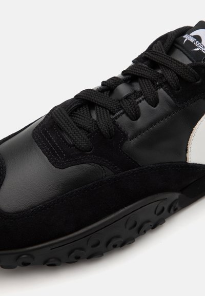 Svart sneaker med en kombination av läder och mocka, texturerade snören och en distinkt gummisula med rundade detaljer.