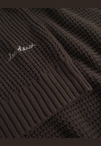 Brown knitted blanket with textured waffle and rib patterns, featuring subtle white embroidered text reading "les deux." Soft, chunky material.