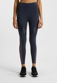 Woman wearing navy blue leggings with sheer panel details and black sneakers, standing against a plain light background.