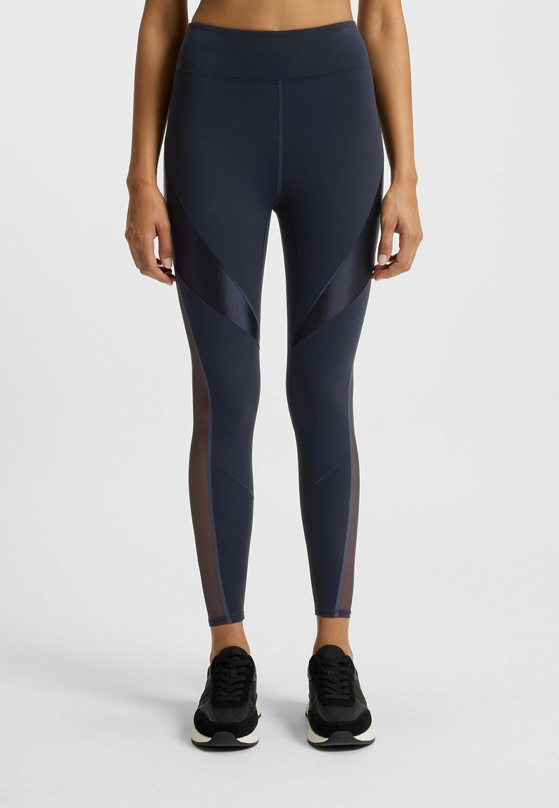 Woman wearing navy blue leggings with sheer panel details and black sneakers, standing against a plain light background.