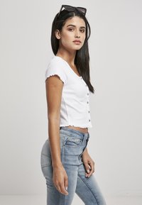 Biele pruhované crop top s okrúhlym výstrihom a volánovými rukávmi, so zapínaním na prednej strane; skombinovaný s svetlomodrými džínsami s vysokým pásom.