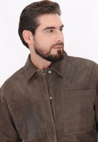 Veste chemise en cuir marron avec un col, des boutons-pression en argent et deux poches avant. Texture mate, détails de couture minimalistes.