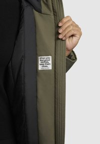 Olijfgroene jas met zwarte voering, met een zichtbaar label met tekst. De stof is soepel, met gestikte details en een zichtbare pocket.