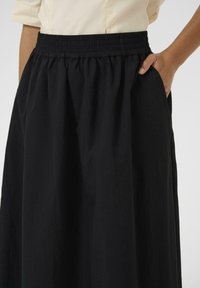 Fekete, elasztikus derékú szoknyát viselő személy, akinek a keze részben a zsebben van, és bézs felsőt visel, ami a derék fölött végződik.