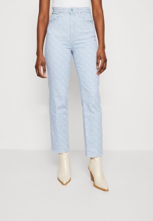 Persoon draagt lichtblauwe jeans met hoge taille en subtiel patroon en witte puntige enkellaarzen, staande tegen een effen achtergrond.
