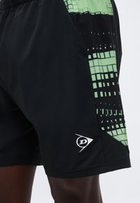 Μαύρο αθλητικό σορτς με πράσινο γεωμετρικό σχέδιο στο πλάι και μικρό λευκό λογότυπο "D" κοντά στο τελείωμα, που φοριέται σε σκουρόχρωμο πόδι.