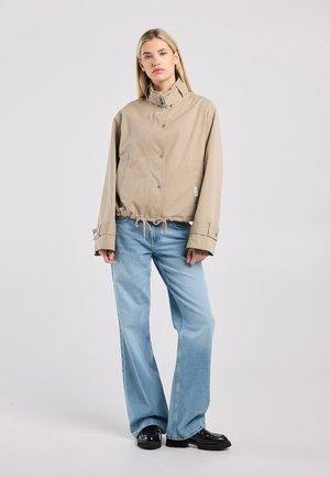 Femme aux longs cheveux blonds portant une veste beige boutonnée, un jean large bleu clair et des chaussures noires, debout devant un fond clair.