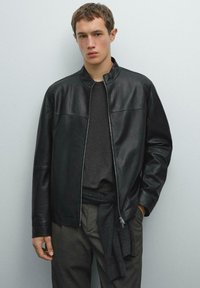 Young man wearing a black leather jacket over a dark shirt, grey pinstripe trousers, and a sweater tied around his waist, standing against a plain wall.