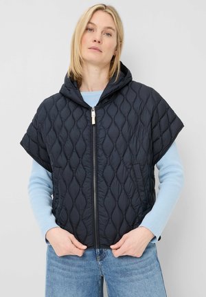 Blonde Frau trägt eine schwarze gesteppte kurzärmelige Kapuzenweste über einem hellblauen Pullover und blauen Jeans, steht mit den Händen nahe dem Reißverschluss.