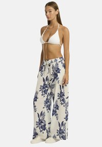 La modella è in piedi di lato indossando un top bikini bianco e pantaloni larghi a fiori, con capelli lunghi e lisci e un'espressione neutra.
