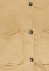 Katoenen vest in lichtbeige met een knoopsluiting aan de voorkant, voorzien van donkere knopen, twee grote opgestikte zakken en stiksels langs de naden.