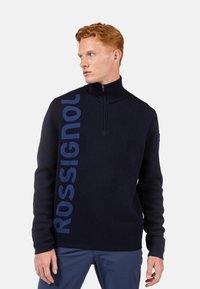 Dunkelblauer Zip-Pullover mit geripptem Kragen, langen Ärmeln und großem "ROSSIGNOL"-Logo in Blau auf der linken Seite.
