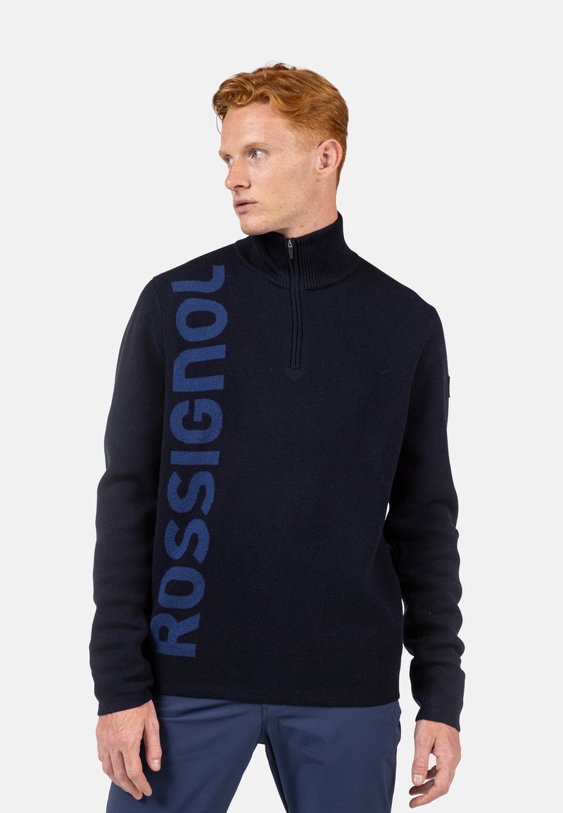 Dunkelblauer Zip-Pullover mit geripptem Kragen, langen Ärmeln und großem "ROSSIGNOL"-Logo in Blau auf der linken Seite.