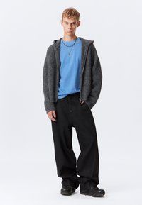 Joven con cabello corto que lleva una camiseta azul, un suéter gris con cremallera, pantalones negros holgados, zapatos negros y collares en capas, de pie contra un fondo blanco.