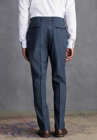 Maßgeschneiderte dunkelblaue Hose mit strukturiertem Finish, ausgestattet mit zwei Gesäßtaschen mit Knopfverschluss und schlankem Schnitt. Sichtbare braune Schuhe.