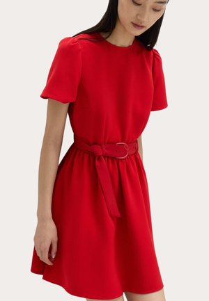 Kobieta w czerwonej sukience z krótkim rękawem, zebranej w talii i z dopasowanym paskiem, stoi na jednolitym jasnym tle.