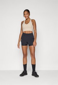 Lys beige sports-bh kombinert med svarte shorts med lagdelt design. Svarte treningssokker og sko fullfører antrekket.