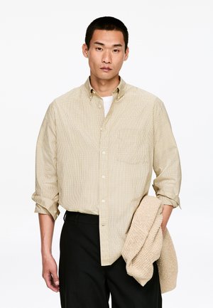 Kariertes Hemd in Beige und Braun mit Knopfleiste, langärmlig, Ärmel hochgekrempelt, kombiniert mit schwarzer Hose, hält einen strukturierten beigen Pullover.