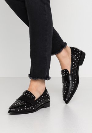 Sorte læderloafers med nitter båret med sorte jeans med frynset kant, fødder krydset på en simpel lys grå gulv og baggrund.