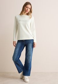 Crèmefarbener Sweatshirt mit langen Ärmeln und "ENDLESS"-Aufdruck, kombiniert mit blauen weit geschnittenen Jeans. Weiße Sneakers runden das Outfit ab.
