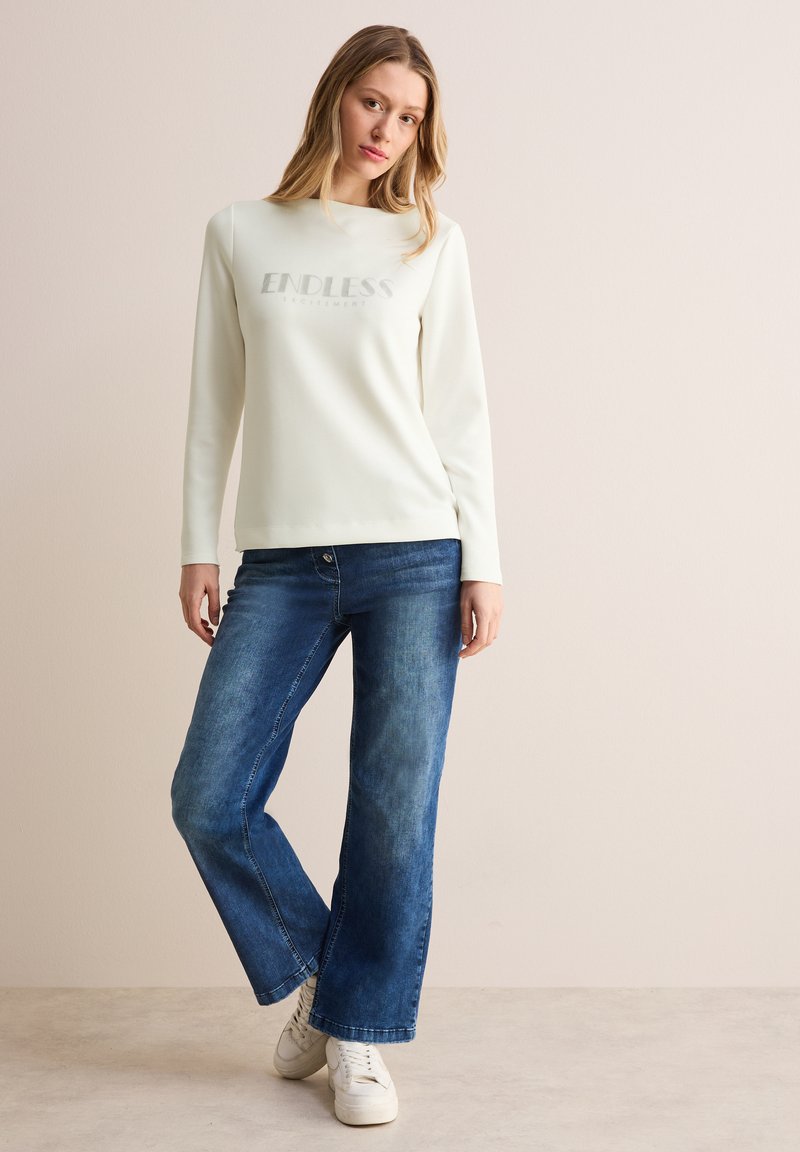 Crèmefarbener Sweatshirt mit langen Ärmeln und "ENDLESS"-Aufdruck, kombiniert mit blauen weit geschnittenen Jeans. Weiße Sneakers runden das Outfit ab.