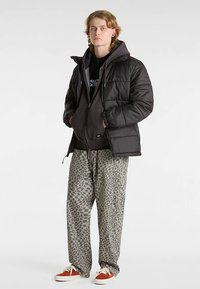 Zwarte donsjas over een donkere hoodie, gecombineerd met grijze, loszittende broek met patroon en oranje-witte sneakers.