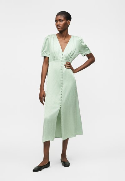 Γυναίκα με κοντά μαλλιά σε ανοιχτοπράσινο μίντι φόρεμα με φουσκωτά μανίκια και κουμπιά καλυμμένα με ύφασμα μπροστά, στέκεται με το ένα χέρι στη μέση, μαύρα επίπεδα παπούτσια.