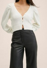 Bílý zkrácený svetr s nafouknutými rukávy a zapínáním na knoflíky, kombinovaný s černými koženými kalhotami s vysokým pasem, hladkou texturou a přiléhavým střihem.